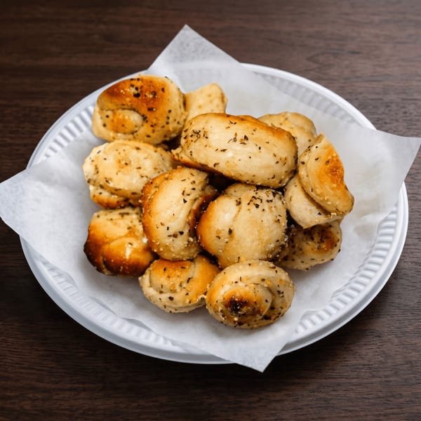 A white plate filled with golden-brown garlic knots, sprinkled with herbs and salt, on a dark wooden table.